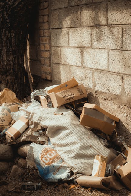 Two large black trash bags made of shiny plastic material are placed on the pavement beside a curb in an outdoor urban environment. The bags appear voluminous and are partly crumpled, with visible cre
